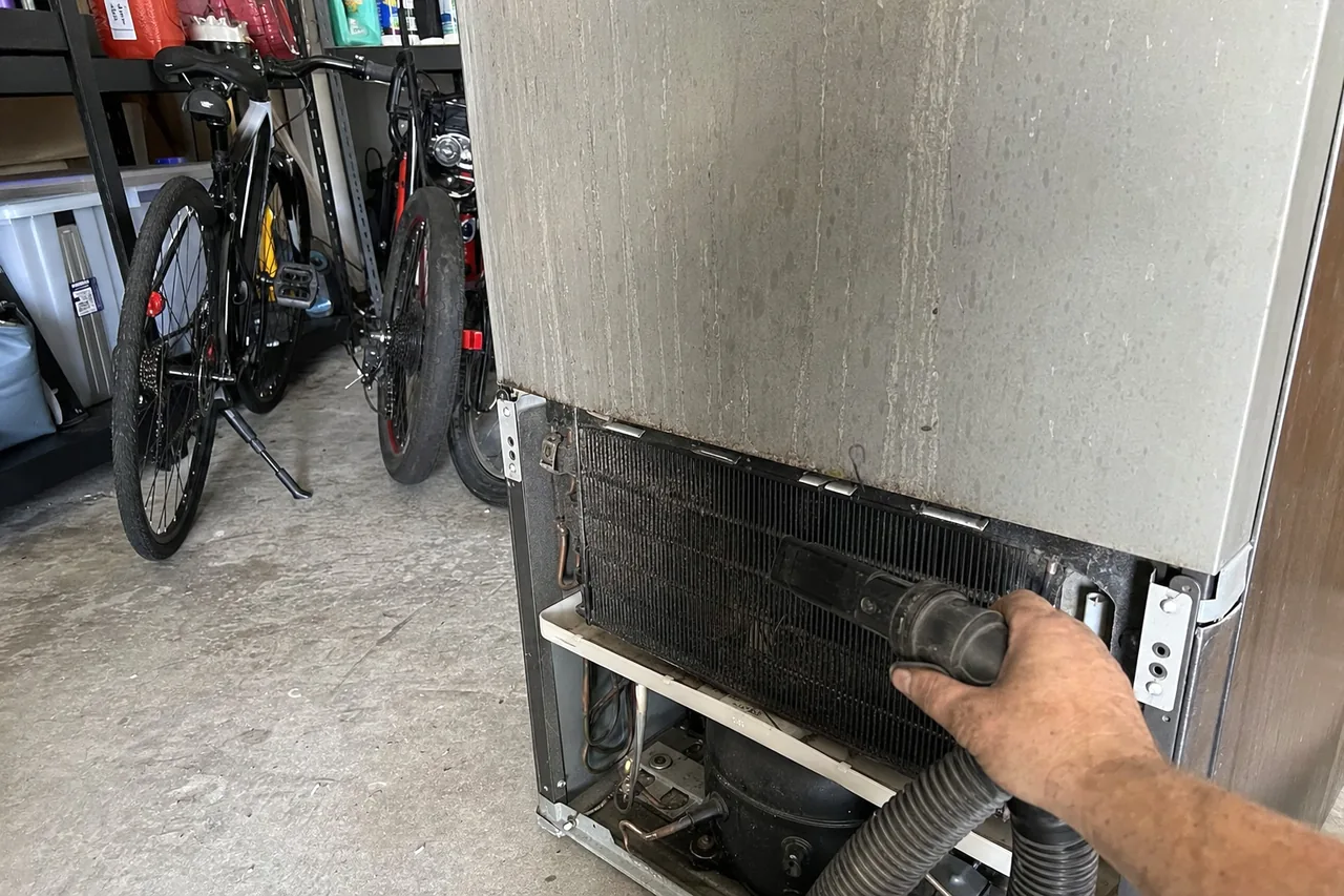 A refrigerator with its lower grille removed for a condenser clean, seen from across a garage floor.
