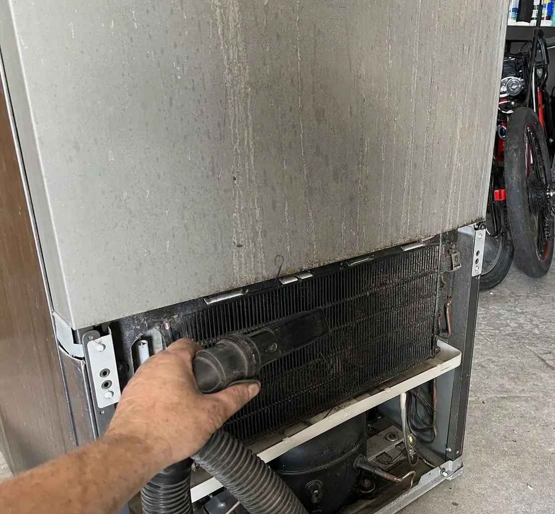 A technician's hand running a vacuum nozzle along the condenser coil behind the lower grille of a refrigerator.