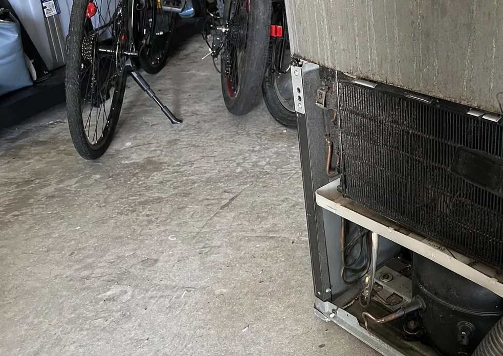 The machine compartment behind a refrigerator grille, compressor mounts and tubing running across the base pan.