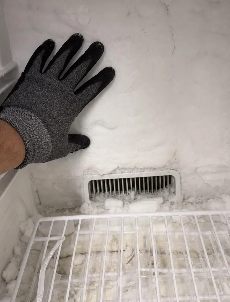A gloved hand pressed to a frosted freezer wall while the vent grille below it stays crusted with ice.